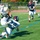 An American football game is in progress on a green turf field with blue markings. Two players are engaged in a tackle or catch attempt near the center of the frame, while a third player wearing number 11 in a navy uniform runs toward the play. A fourth player wearing number 3 is visible on the sideline near the bleachers.