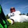 A football player in full uniform is sitting on a grass field, photographed from a low ground-level angle. The dramatic perspective places a bright green Nike cleat in the extreme foreground, with the player's white uniform and red-faced Riddell helmet visible in the background. The image appears to be a stylized portrait or promotional shot taken on a sunny day.