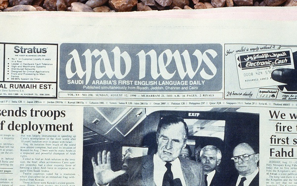 A photograph of the Arab News newspaper front page dated Sunday, August 12, 1990, Saudi Arabia's first English language daily. The front page covers major stories related to the Gulf Crisis following Iraq's invasion of Kuwait, including Egypt sending troops, President Bush's statements, and King Fahd's declaration. The newspaper is laid on a gravel or pebble surface.