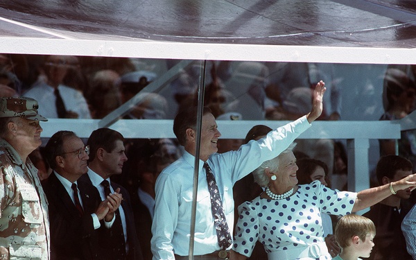 A group of dignitaries and officials are gathered in what appears to be a secure viewing box or reviewing stand, likely at a military parade or public ceremony. One man in a light blue shirt and tie is waving to the crowd while a woman in a white polka-dot dress points outward. A military officer in desert camouflage uniform stands to the left, and a large crowd is visible in the background.