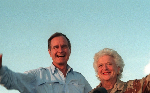 Two older individuals, a man and a woman, stand together at a wooden podium with microphones, both smiling and waving with arms outstretched. An American flag is visible in the background on a flagpole. The setting appears to be an outdoor public or official event, likely during daytime.
