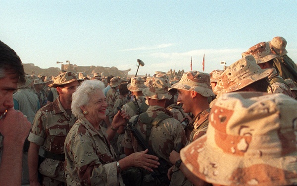 A civilian woman with white hair, wearing desert camouflage clothing, is interacting warmly with a large group of military personnel in desert camouflage uniforms. The setting appears to be an outdoor military gathering in an arid environment, likely during the Gulf War era based on the distinctive 'chocolate chip' desert camouflage pattern. A civilian man in a tan vest is visible on the left side of the frame.