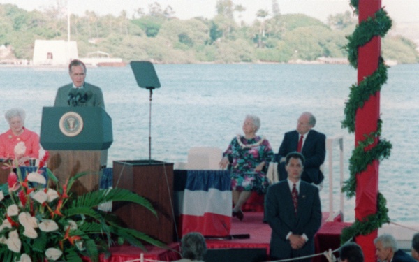 An outdoor official ceremony is taking place at a waterfront location, with a speaker at a podium bearing the Presidential Seal of the United States. The stage is decorated with red, white, and blue bunting, tropical floral arrangements, and a red pole wrapped in green garland. A seated audience faces the stage, and several dignitaries and security personnel are visible on and around the platform.