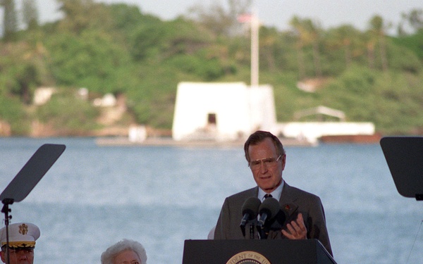 A formal outdoor ceremony is taking place at what appears to be Pearl Harbor, Hawaii, with a speaker at a Presidential podium. A white structure visible across the water in the background appears to be the USS Arizona Memorial. Two individuals are seated behind the speaker — one in a white military dress uniform and one in a coral/pink outfit.