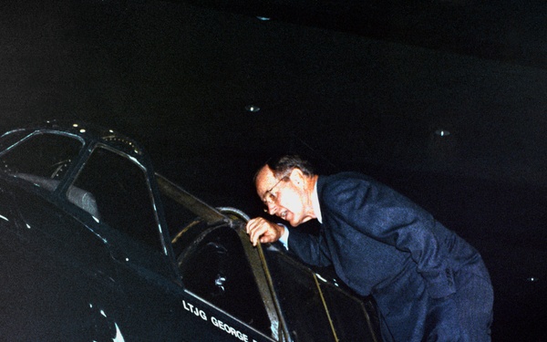 A man in a dark business suit and glasses is leaning into the cockpit of a dark-colored military aircraft. The aircraft bears the inscription 'LTJG GEORGE BUSH USNR' on its fuselage, identifying it as a plane associated with Lieutenant Junior Grade George Bush of the United States Naval Reserve. The image appears to be taken in a museum or exhibition setting given the dark background.