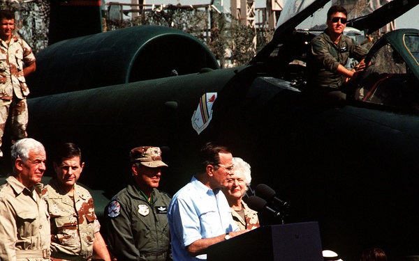 An outdoor military event featuring a civilian man and woman speaking at a podium with a microphone, set against a large dark military aircraft. The crowd includes a mix of military personnel in desert camouflage uniforms, flight suits, and civilians, along with media photographers. The setting appears to be a military air base, likely in a Middle Eastern region based on the uniforms and attendees.