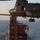 MARSOC conducts VBSS training with 160th SOAR A military special operations fast-rope insertion exercise is underway onto the deck of a container ship at sea. A CH-47 Chinook helicopter hovers above while multiple armed personnel rappel or have already landed on the vessel's deck. The ship bears stacked red shipping containers marked with 'APL' branding and the word 'BREACH' written in green.