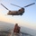 MARSOC conducts VBSS training with 160th SOAR A military helicopter, identified as a CH-47 Chinook tandem-rotor helicopter, hovers over the deck of a large vessel at sea during what appears to be a maritime boarding or fast-rope insertion exercise. Multiple armed personnel in tactical gear are visible on the ship's deck, having apparently fast-roped or rappelled down from the helicopter. Shipping containers marked with 'APL' branding are stacked on the vessel's deck, suggesting this may be a training exercise simulating a vessel interdiction operation.