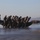 MARSOC conducts VBSS training with 160th SOAR A group of military personnel in camouflage uniforms and helmets are carrying inflatable rubber boats (IBS) across a wet beach. At least two teams are visible, each carrying a large black inflatable boat on their heads or shoulders. The setting appears to be a coastal training environment, consistent with amphibious or special operations training exercises.