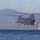 MARSOC conducts VBSS training with 160th SOAR A military CH-47 Chinook tandem-rotor helicopter hovers low over open water near a coastal area with rocky cliffs and mountains in the background. Personnel can be seen hanging or rappelling from the rear of the aircraft, suggesting a special operations water insertion or training exercise. The scene appears to be a maritime helicast or fast-rope operation conducted over the sea.
