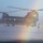 MARSOC conducts VBSS training with 160th SOAR A military CH-47 Chinook helicopter hovers low over water in misty conditions, with two personnel suspended below it on ropes or cables. The scene appears to depict a special operations fast-rope or rappel insertion exercise over a body of water. A rainbow-like light refraction is visible in the mist created by the rotor wash.