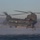 MARSOC conducts VBSS training with 160th SOAR A military CH-47 Chinook helicopter hovers low over open water near a rocky coastline, with rotor wash creating a visible mist/spray over the water surface. A person in tactical gear is descending via a rope ladder from the rear of the aircraft. This appears to be a special operations maritime insertion or training exercise.