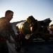 A U.S. Air Force airman in ABU (Airman Battle Uniform) is working with inert training munitions on a weapons loading cart at what appears to be a desert airbase during sunrise or sunset. The munitions are clearly marked 'DO NOT FLY' and 'INERT LOAD CREW,' indicating these are non-live training ordnance used for weapons loading practice. The dramatic backlighting from the low sun creates a strong lens flare effect in the image.