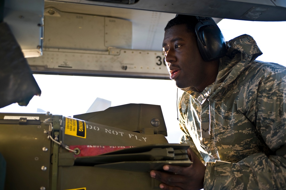 A U.S. Air Force airman in digital camouflage (ABU pattern) uniform and hearing protection headset is working on or inspecting ordnance/munitions mounted under an aircraft. A 'DO NOT FLY' warning label is clearly visible on the munition, along with a yellow 'WARNING' sticker. The image is taken from a close, low-angle perspective beneath the aircraft fuselage.