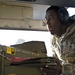 A U.S. Air Force airman in digital camouflage (ABU pattern) uniform and hearing protection headset is working on or inspecting ordnance/munitions mounted under an aircraft. A 'DO NOT FLY' warning label is clearly visible on the munition, along with a yellow 'WARNING' sticker. The image is taken from a close, low-angle perspective beneath the aircraft fuselage.