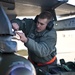 A military airman, identifiable by his olive green flight suit and rank insignia, is working on ordnance mounted under the wing of a military aircraft. He is wearing hearing protection ear muffs and an orange safety vest, indicating active flight line operations. The scene appears to be on a military airfield tarmac during daylight hours.