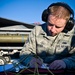 A U.S. Air Force airman wearing digital camouflage ABU (Airman Battle Uniform) and hearing protection is writing on a clipboard or notepad on a flight line. The individual, whose name tape reads 'OTTER' (partially visible), is positioned in front of a military jet aircraft loaded with what appears to be a munition on its wing pylon. The image appears to be an official military documentation or maintenance activity photograph.