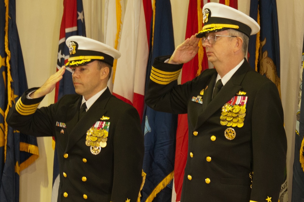 Two U.S. Navy officers in dress blue uniforms are rendering a salute during a formal military ceremony. Both wear white-topped service caps with gold insignia and display extensive medal and ribbon arrays on their chests. The backdrop consists of multiple U.S. state and military flags with gold fringe, indicating an official indoor ceremony.
