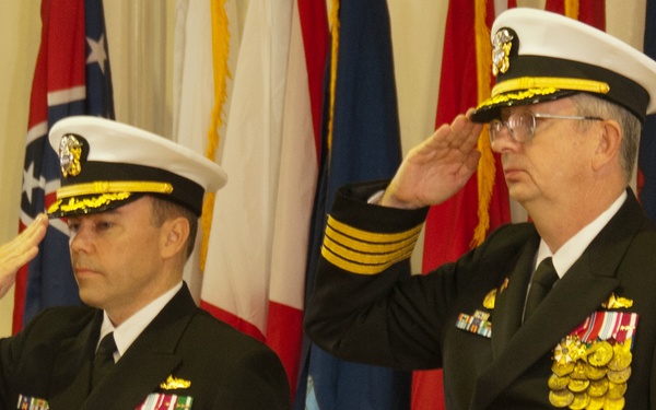 Two U.S. Navy officers in dress blue uniforms are rendering a salute during a formal military ceremony. Both wear white-topped service caps with gold insignia and display extensive medal and ribbon arrays on their chests. The backdrop consists of multiple U.S. state and military flags with gold fringe, indicating an official indoor ceremony.