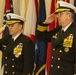 Two U.S. Navy officers in dress blue uniforms are rendering a salute during a formal military ceremony. Both wear white-topped service caps with gold insignia and display extensive medal and ribbon arrays on their chests. The backdrop consists of multiple U.S. state and military flags with gold fringe, indicating an official indoor ceremony.
