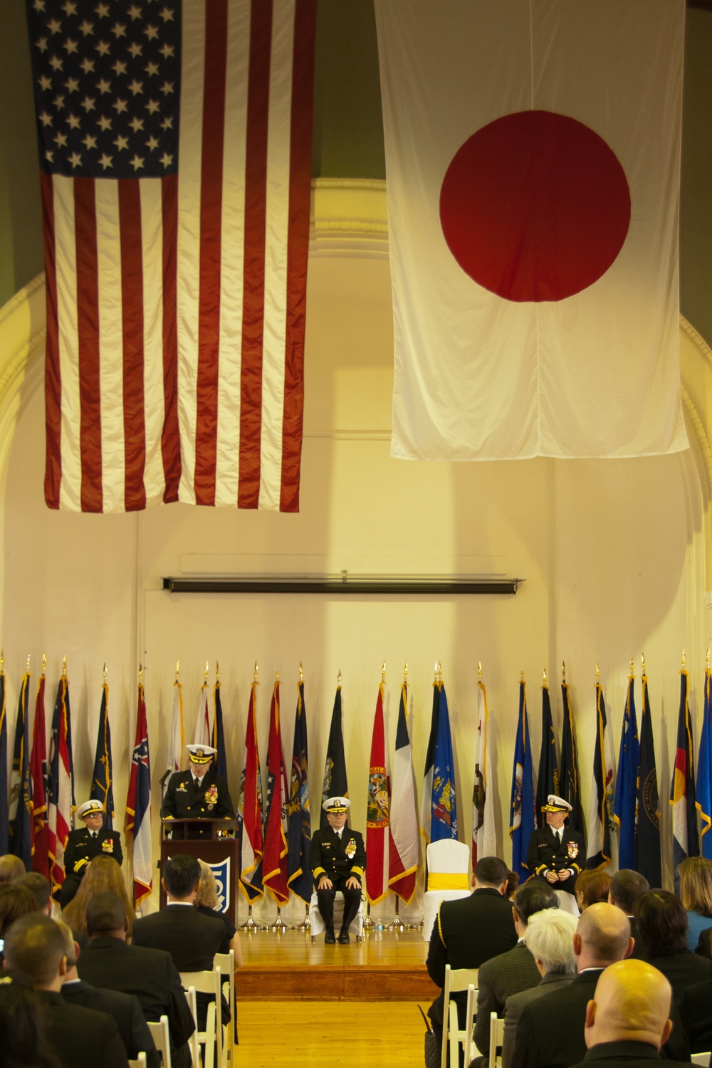 A formal military ceremony is taking place in a large ceremonial hall, with large flags of the United States and Japan prominently displayed overhead. Several naval officers in dress uniforms are positioned on a stage, with one speaking at a podium, while an audience seated in white chairs observes. A row of what appear to be U.S. state flags lines the back of the stage.