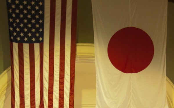 A formal military ceremony is taking place in a large ceremonial hall, with large flags of the United States and Japan prominently displayed overhead. Several naval officers in dress uniforms are positioned on a stage, with one speaking at a podium, while an audience seated in white chairs observes. A row of what appear to be U.S. state flags lines the back of the stage.