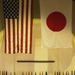 A formal military ceremony is taking place in a large ceremonial hall, with large flags of the United States and Japan prominently displayed overhead. Several naval officers in dress uniforms are positioned on a stage, with one speaking at a podium, while an audience seated in white chairs observes. A row of what appear to be U.S. state flags lines the back of the stage.