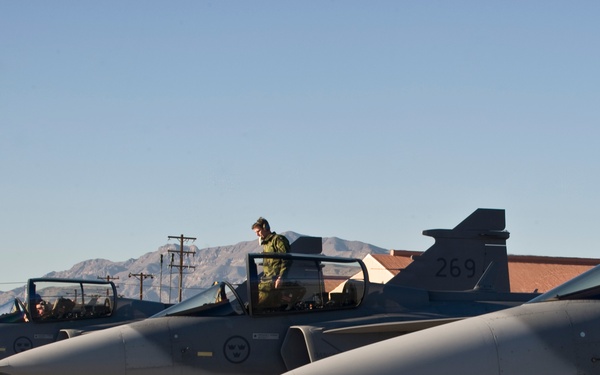 A row of Swedish Air Force Saab JAS 39 Gripen fighter jets are lined up on a military airfield tarmac, likely at Nellis Air Force Base in Nevada based on the desert mountain backdrop. Multiple pilots and ground crew in olive/green flight suits are visible in and around the cockpits, suggesting pre-flight or post-flight operations. The aircraft bear Swedish Air Force roundel markings and at least one visible tail number '269'.