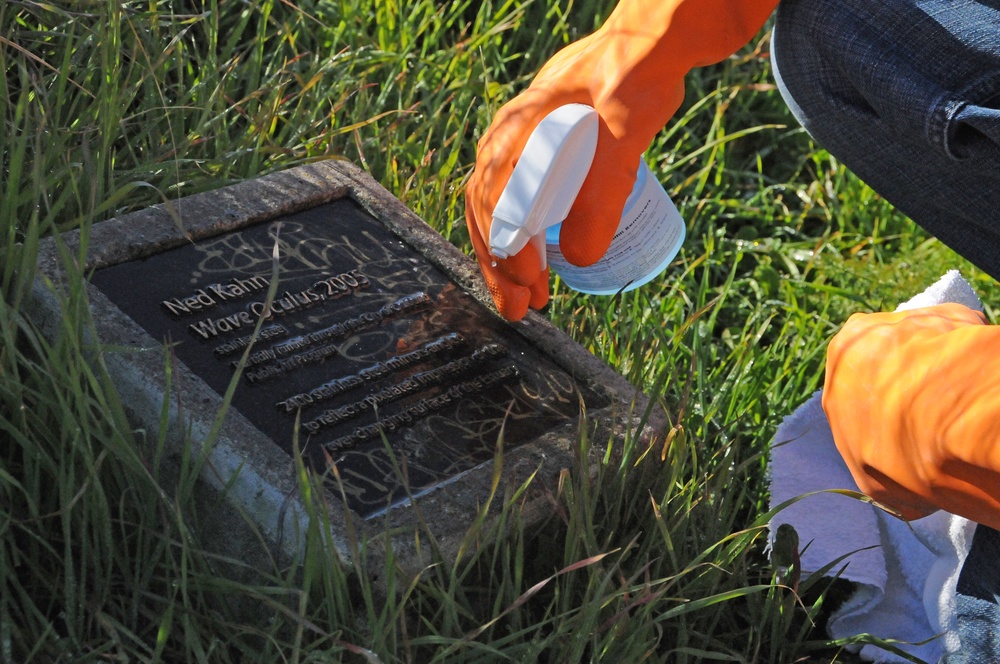 A person wearing orange rubber gloves is cleaning a vandalized commemorative plaque using a spray bottle and cloth. The dark stone plaque, embedded in a grassy area, bears the name 'Ned Kahn' and references 'Wave Oculus, 2005' but is covered in graffiti. The cleaning activity appears to be graffiti removal from a public art installation marker.