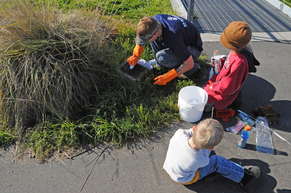 An overhead view of three people — one adult and two children — engaged in what appears to be an outdoor cleaning or environmental maintenance activity near a grassy area and pavement. The adult is wearing orange rubber gloves and working with a dark container near overgrown grass, while the children sit nearby on the pavement. Various supplies including a white bucket, bottles, and bags are visible on the ground.
