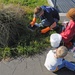 An overhead view of three people — one adult and two children — engaged in what appears to be an outdoor cleaning or environmental maintenance activity near a grassy area and pavement. The adult is wearing orange rubber gloves and working with a dark container near overgrown grass, while the children sit nearby on the pavement. Various supplies including a white bucket, bottles, and bags are visible on the ground.