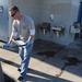 A man wearing blue latex gloves and sunglasses is carrying a black tray or grate near an outdoor public restroom facility. He appears to be performing maintenance or cleanup work, as evidenced by his protective gloves and the dark wet stains visible on the concrete ground. The setting is an outdoor public facility with wall-mounted sinks and a drinking fountain.