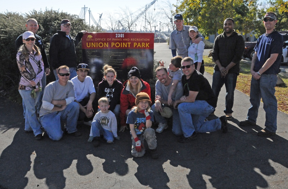 A group of approximately 16-18 people, including adults and two young children, pose together in front of the Union Point Park sign at 2301, managed by the Office of Parks and Recreation. The group appears to be volunteers or community members gathered for a park event or cleanup day, as evidenced by work gloves visible on at least one child. The photo is taken outdoors on a sunny day with trees and industrial structures visible in the background.