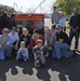 A group of approximately 16-18 people, including adults and two young children, pose together in front of the Union Point Park sign at 2301, managed by the Office of Parks and Recreation. The group appears to be volunteers or community members gathered for a park event or cleanup day, as evidenced by work gloves visible on at least one child. The photo is taken outdoors on a sunny day with trees and industrial structures visible in the background.
