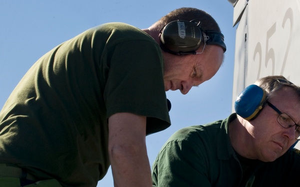 Two military maintenance technicians are working on the surface of an aircraft, visible by the tail number '252' in the background. Both men are wearing green military-style clothing and hearing protection (ear defenders), with one also wearing glasses and teal/green protective gloves. They appear to be performing inspection or repair work on the aircraft's exterior panel.