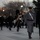 Presidential Inaugural Parade A military parade or ceremonial procession is taking place in an urban setting, likely Washington D.C., during early morning or dusk hours. The foreground features a prominent officer in a gray dress uniform coat with gold buttons and a ceremonial sword, accompanied by another officer in dark dress uniform. Behind them, a military band and marching unit in dark uniforms with white gloves proceed in formation.