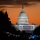 57th Presidential Inauguration The United States Capitol Building is photographed at dusk or dawn, illuminated against a dramatic orange sky. The building is decorated with large American flag banners along its facade, consistent with an inauguration ceremony setup. The foreground shows a crowded, lit boulevard — likely the National Mall — with vehicle lights and street lamps visible.