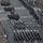 57th Presidential Inauguration An aerial view of a large military parade proceeding down a wide multi-lane urban boulevard. Multiple military units in dark dress uniforms are marching in formation, accompanied by a marching band with visible brass instruments including sousaphones. Spectators line the barricaded sidewalks on both sides of the street.
