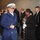 57th Presidential Inauguration A senior military officer in a blue dress uniform with gold buttons and a white peaked cap with gold insignia stands prominently in the foreground, smiling and adjusting black gloves. He is surrounded by a group of people in formal attire, including other military personnel and civilians, in what appears to be an indoor ceremonial or official event setting. A woman in the background holds a ceremonial mace or staff topped with an eagle figure.