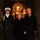 57th Presidential Inauguration Three individuals are posed together in what appears to be a formal indoor entryway or corridor with warm ambient lighting. The group includes a military officer in dress uniform on the left, a woman in a black coat with glasses in the center, and an older man in a black overcoat on the right. A red carpet and blue runner are visible on the floor, suggesting a formal or ceremonial occasion.
