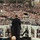 57th Presidential Inauguration A person in a dark suit stands at a podium with teleprompters, addressing an enormous crowd gathered outdoors. The setting features a white balustrade with glass panels and arched architectural details, consistent with a large formal public ceremony. The crowd extends far into the background, with bare winter trees visible in the distance.