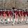 57th Presidential Inauguration A military ceremonial unit dressed in 18th-century Continental Army-style uniforms marches in formation on a paved plaza. The unit appears to be the U.S. Army Old Guard Fife and Drum Corps, identifiable by their distinctive red coats, tricorn hats, and period-accurate regalia. A drum major or commander leads the formation at the front, wearing a tall bearskin cap and carrying a ceremonial sword.