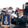 Presidential Inaugural Parade A military musician in formal dress uniform plays a silver trumpet/bugle during an outdoor ceremonial event. A banner attached to the instrument displays the 'Pershing's Own' insignia, identifying the player as a member of the U.S. Army Band. A large crowd of spectators surrounds the performer, many photographing the event with smartphones and cameras.