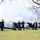 Presidential Inaugural Parade A formal military cannon salute ceremony is taking place outdoors on a grassy area surrounded by bare winter trees. Multiple artillery pieces are being fired simultaneously, with visible muzzle flash and smoke emanating from at least one cannon. Uniformed personnel in dark dress uniforms with peaked caps are stationed at each cannon.
