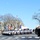 Presidential Inaugural Parade A formal military or government parade is taking place on a wide urban boulevard, likely in Washington D.C., based on the neoclassical architecture visible in the background. A large formation of police or military motorcycles with sidecars is proceeding down the street, accompanied by color guard units carrying multiple flags including the American flag. Uniformed military personnel are standing at attention along the right side of the street in a saluting formation.