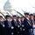 Presidential Inaugural Parade A military honor guard unit is standing in formation in front of the United States Capitol building in Washington, D.C. The personnel are dressed in formal dark navy dress uniforms with white covers (caps) and white gloves, each carrying a rifle with a fixed bayonet at shoulder arms. The scene appears to be a ceremonial parade or inaugural event given the formal attire and Capitol backdrop.