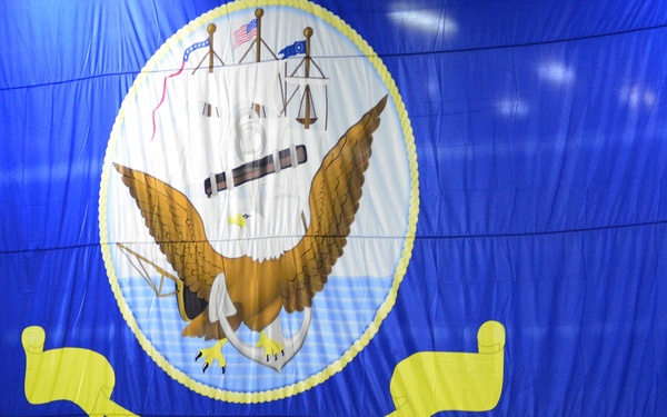 A formal United States Navy ceremony is taking place indoors, with a large assembly of uniformed Navy personnel standing in formation beneath an oversized U.S. Navy seal banner. The banner displays the official Navy emblem featuring a bald eagle, anchor, and ship, with 'UNITED STATES NAVY' written on a yellow ribbon. Portable propane heaters are visible among the assembled sailors, suggesting the event is held in a large hangar or indoor venue during cooler temperatures.