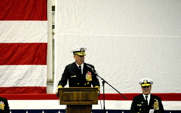 A formal U.S. Navy military ceremony is taking place indoors, with a senior officer speaking at a wooden podium before an audience. Three additional naval officers in dress uniforms are seated on stage behind the speaker, with a large American flag serving as a backdrop. The audience, visible in the foreground, includes personnel in white covers (caps) and civilians.