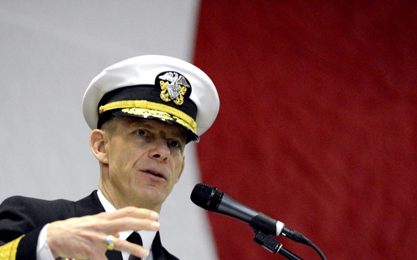 A U.S. Navy officer in dress uniform is delivering a speech at a wooden podium equipped with a microphone. The officer is gesturing with his right hand, suggesting an emphatic or explanatory point during the address. The background features draped fabric in red and white, consistent with a formal military ceremony.
