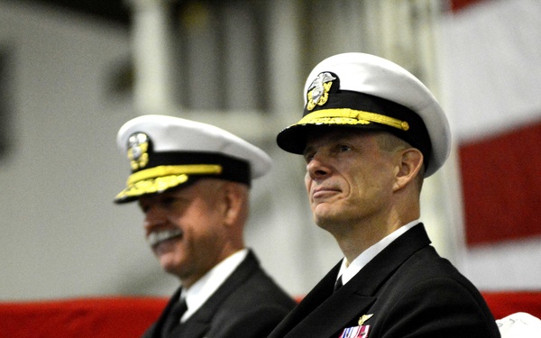 Two U.S. Navy flag officers in dress blue uniforms are seated at what appears to be a formal military ceremony. Both wear white combination covers with gold scrambled eggs insignia, and display extensive medal and ribbon arrays on their chests. An American flag is prominently displayed in the background.